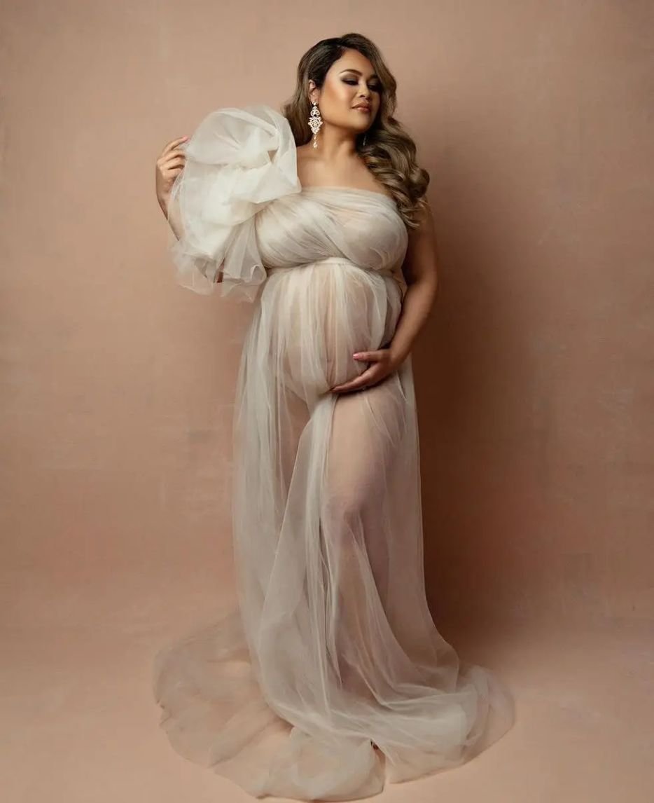 A pregnant woman wears a flowing tulle dress for a maternity photoshoot, highlighting the soft and ethereal quality of the fabric as it drapes elegantly around her.