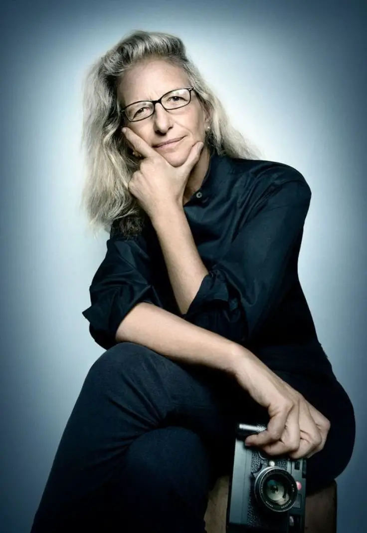 A woman wearing glasses sits on a stool, holding a camera, ready to capture moments.