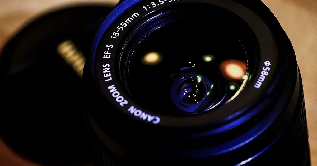 Close-up of a professional camera with a large lens, set on a tripod, capturing a beautifully arranged still life scene featuring various objects like fruits, flowers, and ceramics on a rustic wooden table.