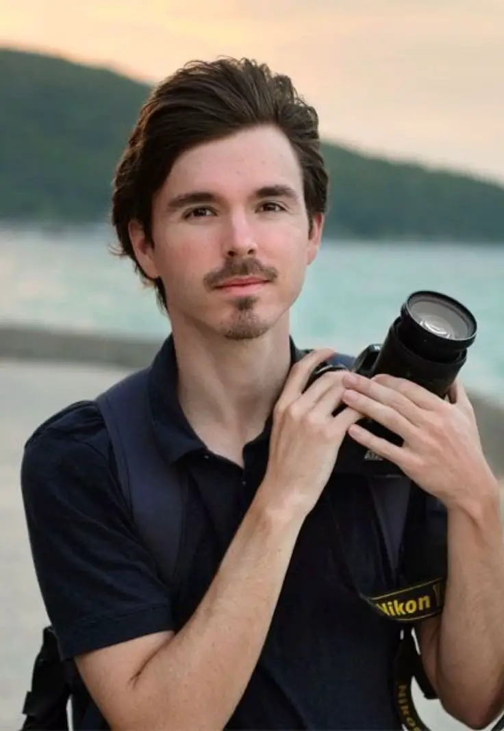 A man stands on the beach, holding a camera, capturing the scenic beauty of the ocean and shoreline.