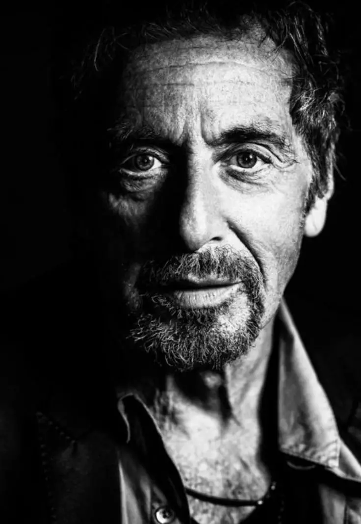 Black and white close-up portrait of a man with a beard and intense eyes, wearing a suit and an open-collared shirt, looking directly at the camera.