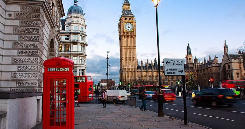 A red phone booth stands prominently on a street corner, surrounded by urban scenery and pedestrian activity.