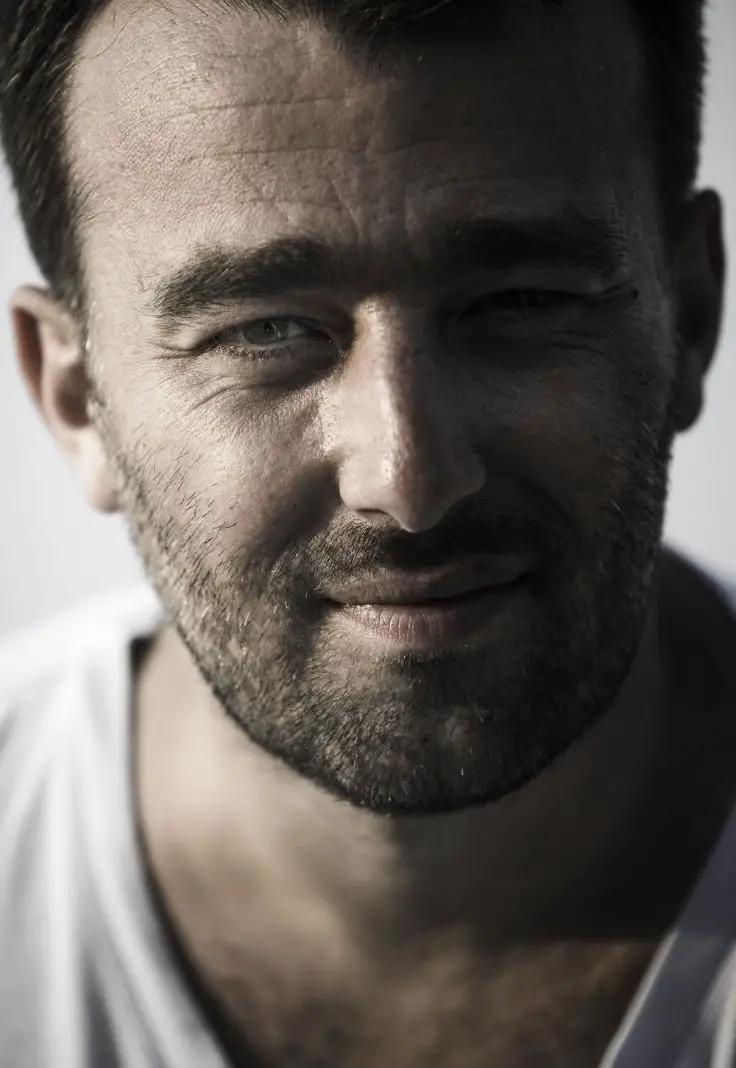 A bearded man wearing a white t-shirt, looking directly at the camera with a neutral expression.