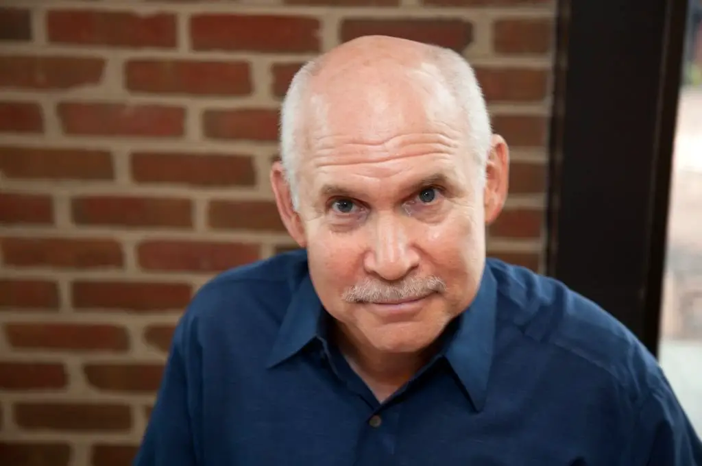 A balding man wearing a blue shirt and sporting a mustache, smiling confidently at the camera.