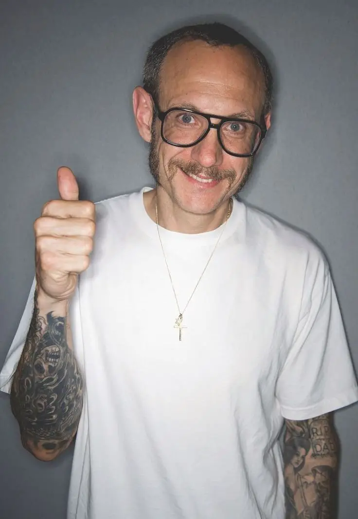 A man with a mustache, sideburns, and large black-framed glasses is smiling and giving a thumbs-up gesture.