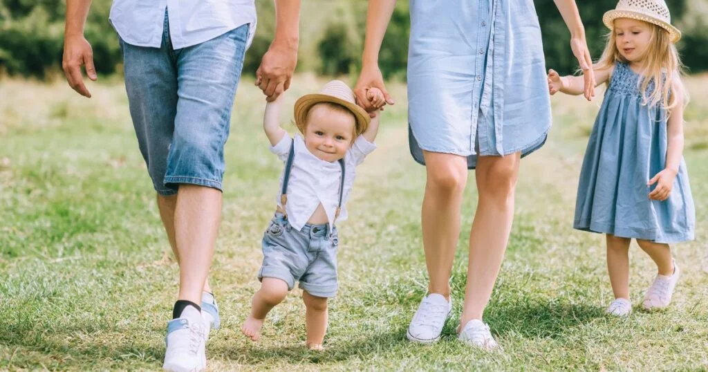 A family dressed in coordinated outfits walks together on lush grass, joyfully carrying their baby.