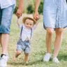 A family dressed in coordinated outfits walks together on lush grass, joyfully carrying their baby.