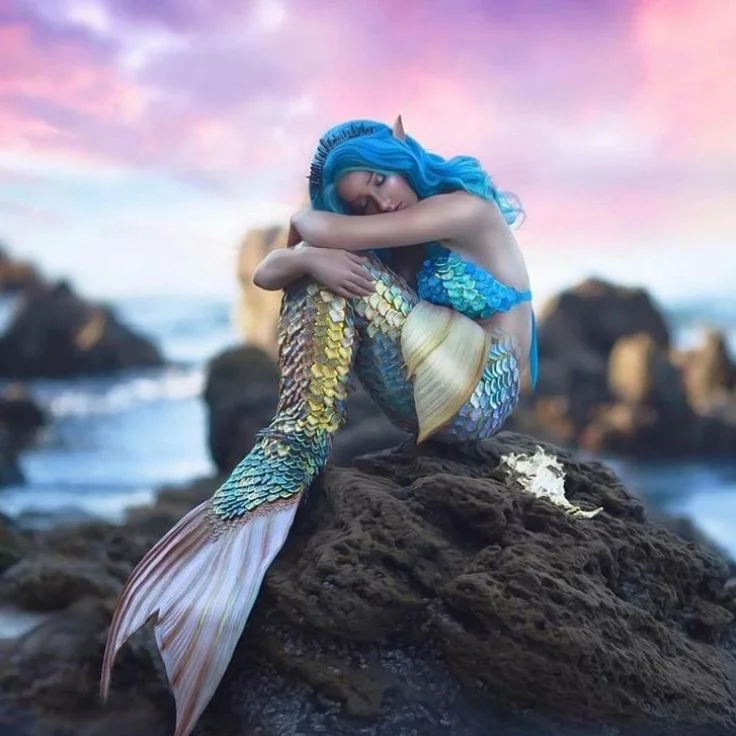 A serene mermaid perched on a rock, arms folded, surrounded by the beauty of the ocean in a captivating photograph.