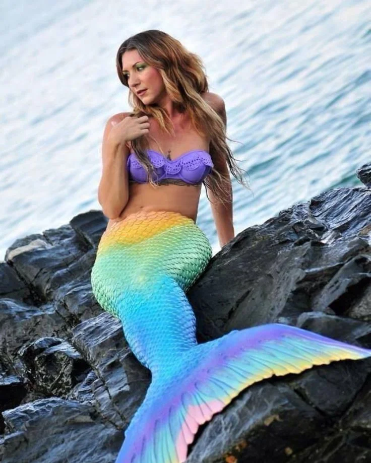 A woman in a vibrant mermaid tail gracefully sits on a rock, embodying the enchanting allure of mermaid photography.