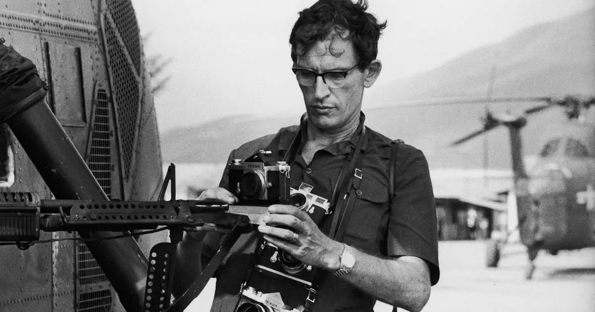 A famous war photographer captures a moment, holding his camera as he gazes at a helicopter overhead